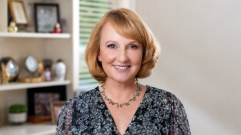 Smiling woman with short blonde hair and blue eyes, wearing a floral blouse and necklace, posed in a softly lit home interior with blurred shelves and framed photos in the background.