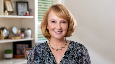 Smiling woman with short blonde hair and blue eyes, wearing a floral blouse and necklace, posed in a softly lit home interior with blurred shelves and framed photos in the background.