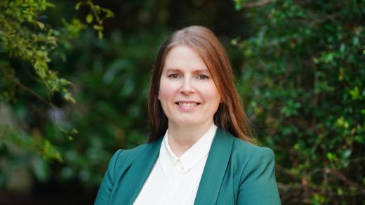 Woman in green sports coat smiling and standing in front of green foliage